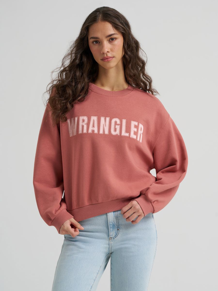 A woman wearing a dusty rose crewneck sweatshirt with 'WRANGLER' printed in white across the chest, paired with light blue jeans, against a light gray studio background.