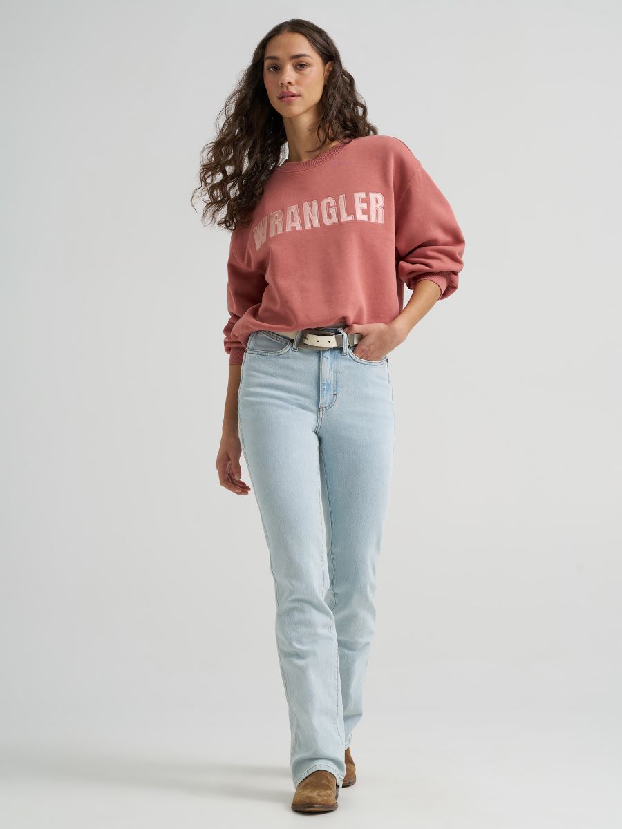 A woman modeling a dusty rose Wrangler sweatshirt with white varsity lettering, paired with light-wash blue jeans and brown suede boots, against a plain light gray studio background.
