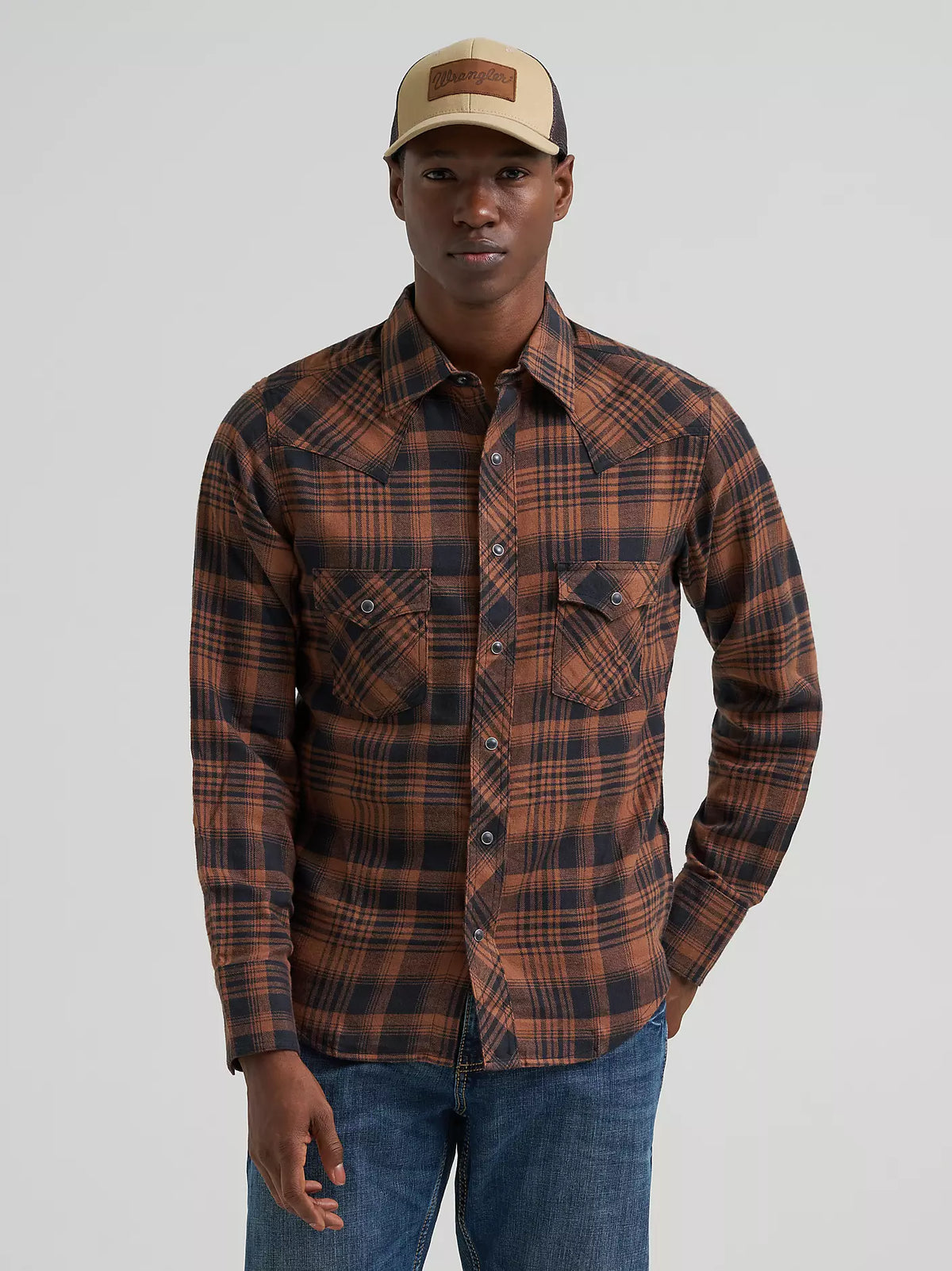 man wearing a brown plaid Wrangler flannel shirt, blue jeans and a ball cap on a grey background