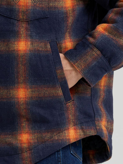 Man wearing a blue and orange Wrangler flannel jacket with his hands in his pocket on a grey background