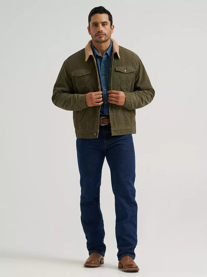 A man modeling an olive green corduroy jacket from Wrangler with a tan shearling collar, worn over a blue button-down shirt and dark blue jeans, standing against a light gray studio background.