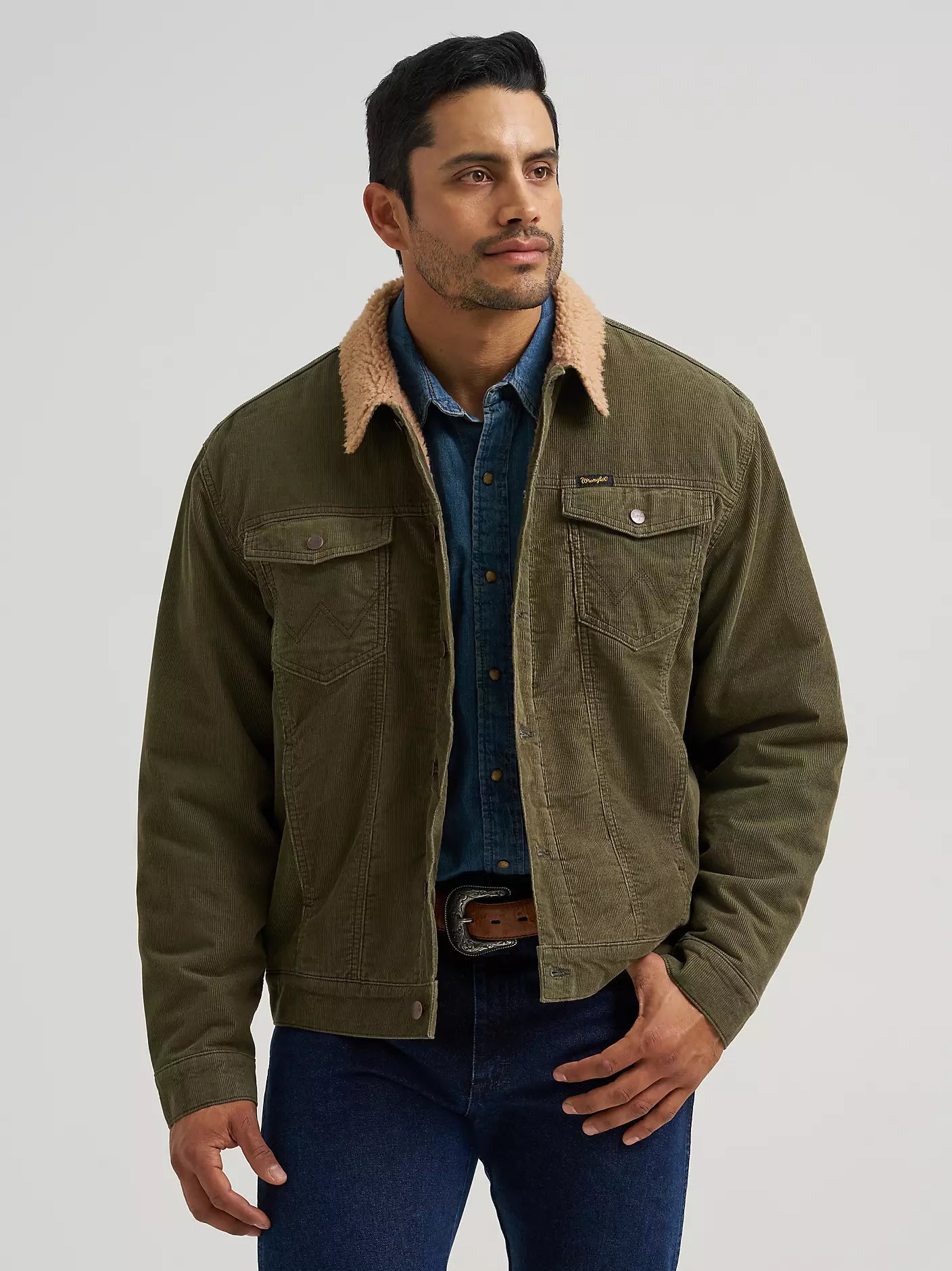 A man wearing an olive green Wrangler corduroy jacket with a tan sherpa collar, layered over a blue denim shirt and jeans, photographed against a light gray studio background.