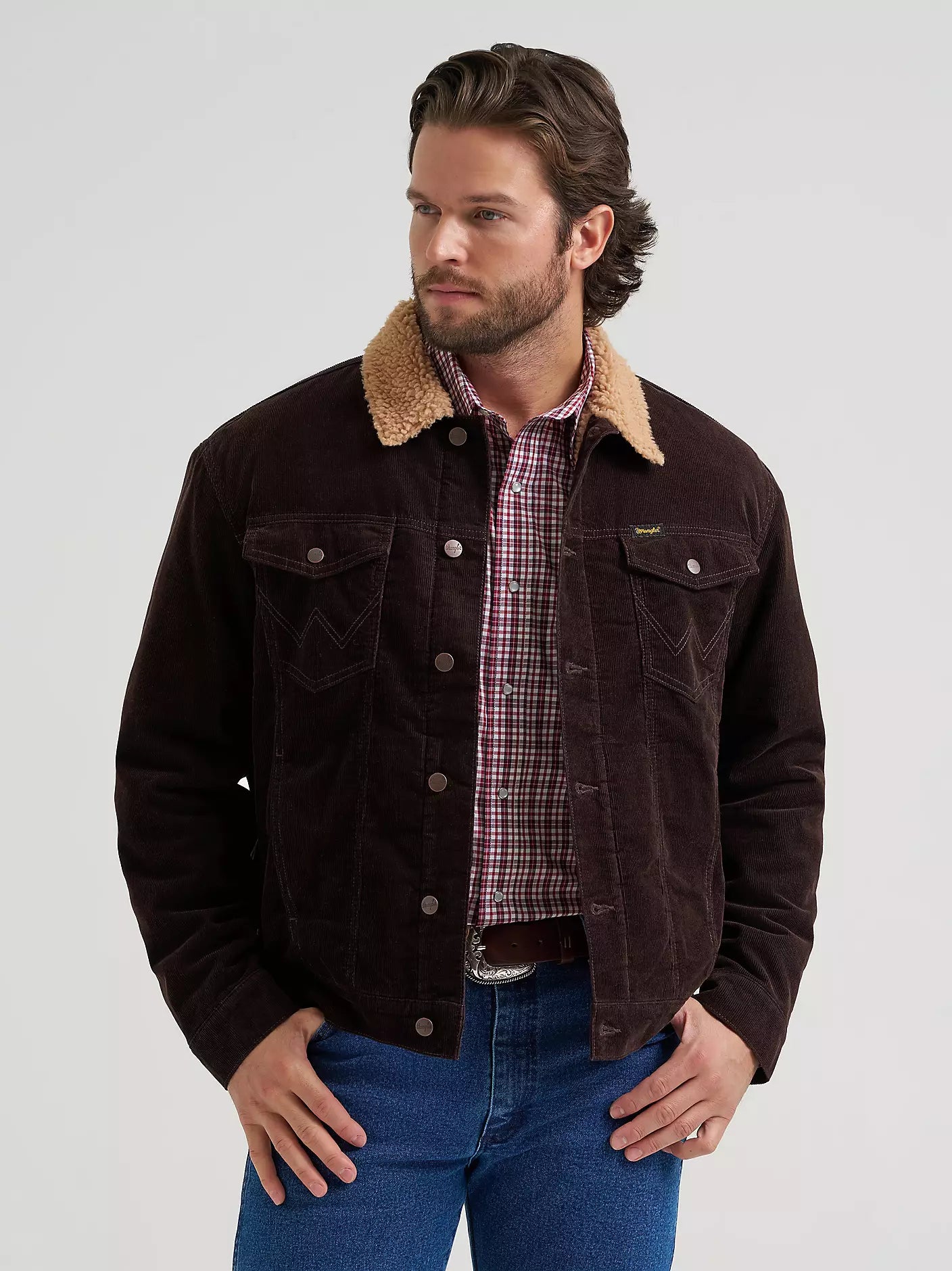 A man wearing a dark brown corduroy Wrangler jacket with a tan sherpa collar, layered over a red and white plaid button-down shirt and blue jeans, photographed against a light gray studio background.