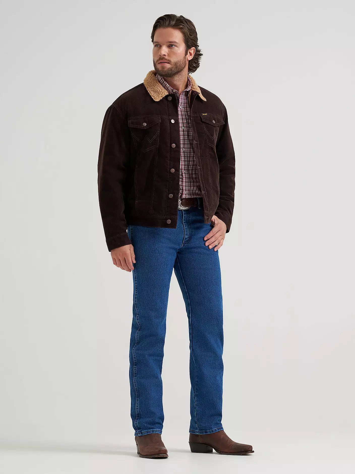 A man wearing a dark brown corduroy Wrangler jacket with a tan corduroy collar, blue jeans, and brown boots, standing against a light gray studio background.