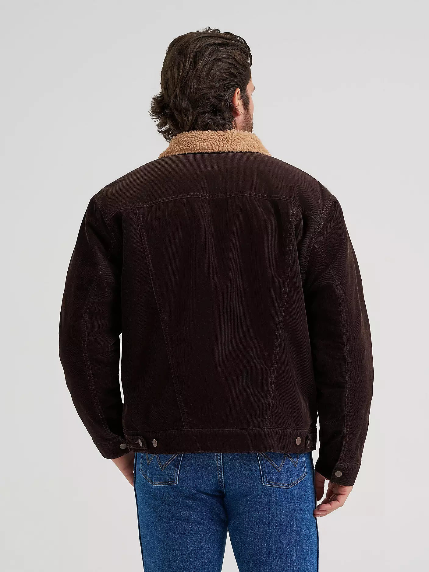 A man viewed from behind wearing a dark brown corduroy Wrangler jacket with a tan sherpa collar and blue jeans, against a light gray studio background.