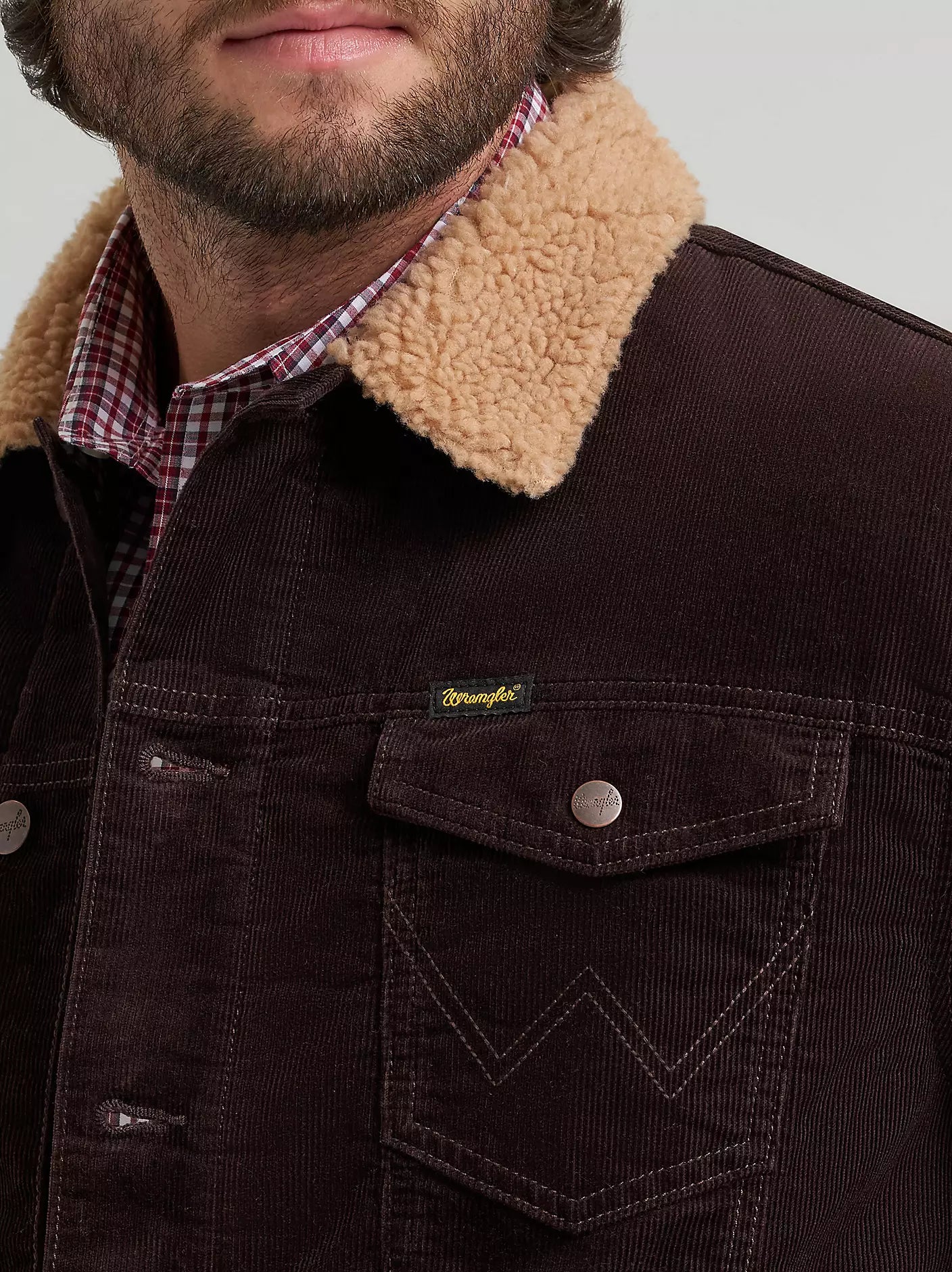 Close-up of a person wearing a dark brown corduroy Wrangler jacket with a tan sherpa collar, layered over a red and white plaid shirt. The Wrangler brand tag is visible on the chest pocket.