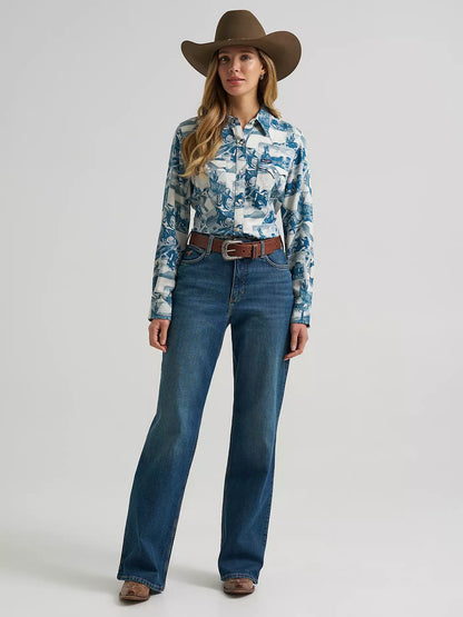 A woman wearing a long-sleeve button-up shirt from Wrangler with a blue and white collage print, paired with blue jeans, shown against a light gray studio background.