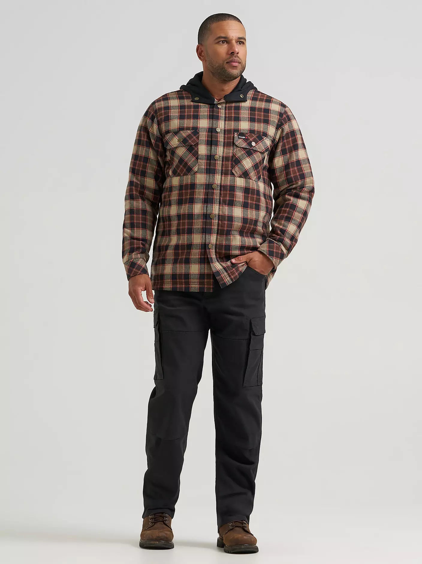 A man wearing a hooded plaid Wrangler jacket with a brown, beige, and black pattern, paired with black cargo pants, photographed against a light gray studio background.