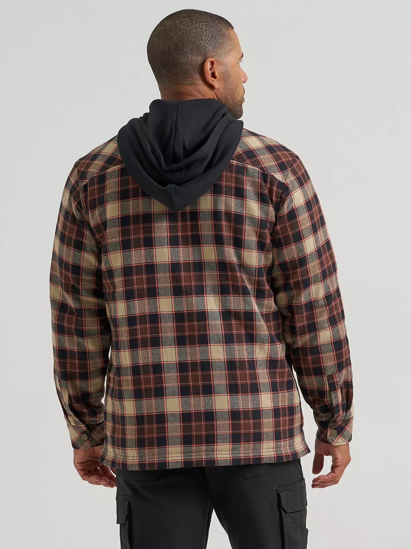 A man wearing a hooded plaid Wrangler jacket with a brown, beige, and black pattern, paired with black cargo pants, photographed against a light gray studio background.