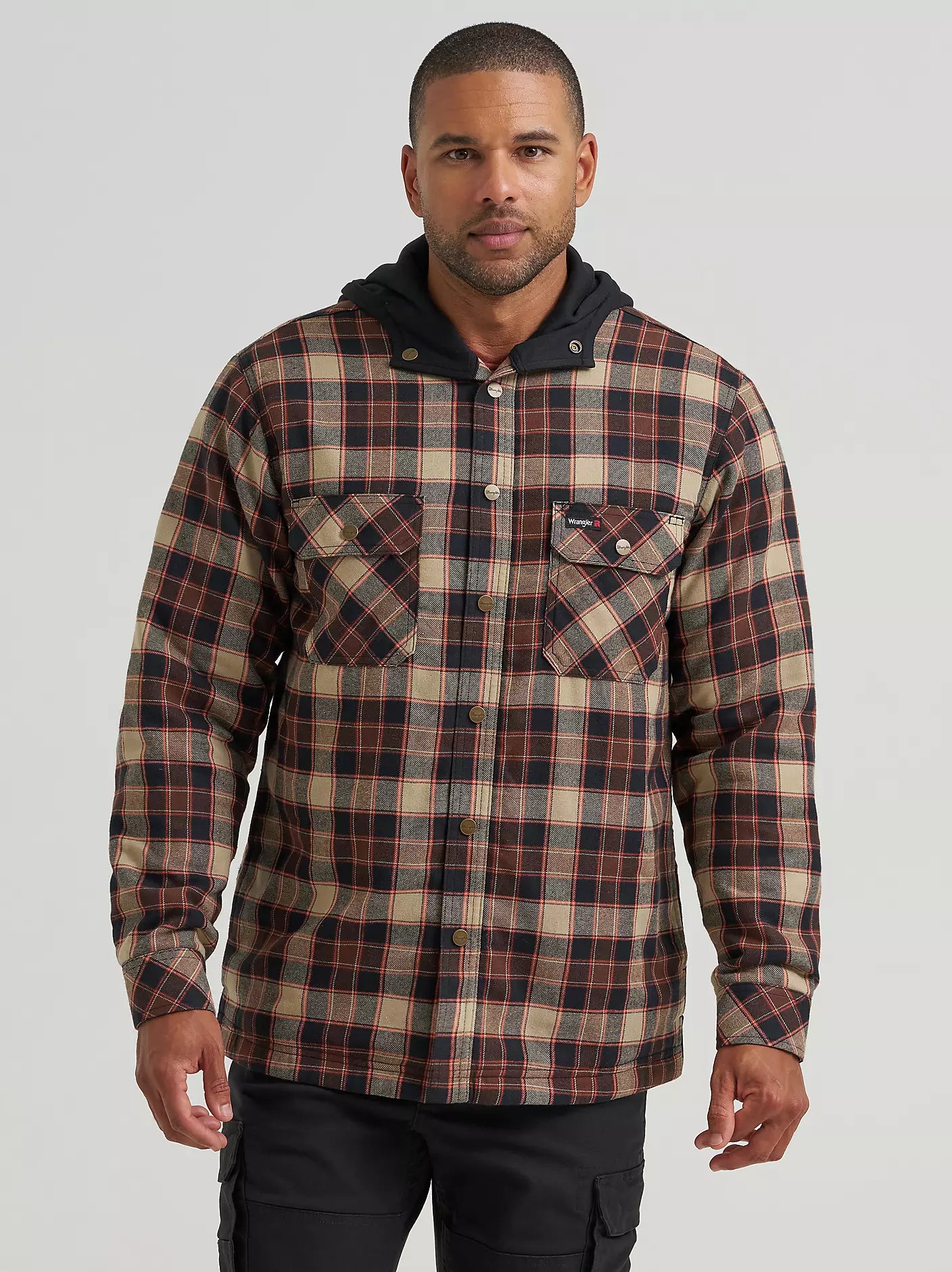A man wearing a hooded plaid Wrangler jacket with a brown, beige, and black pattern, paired with black cargo pants, photographed against a light gray studio background.