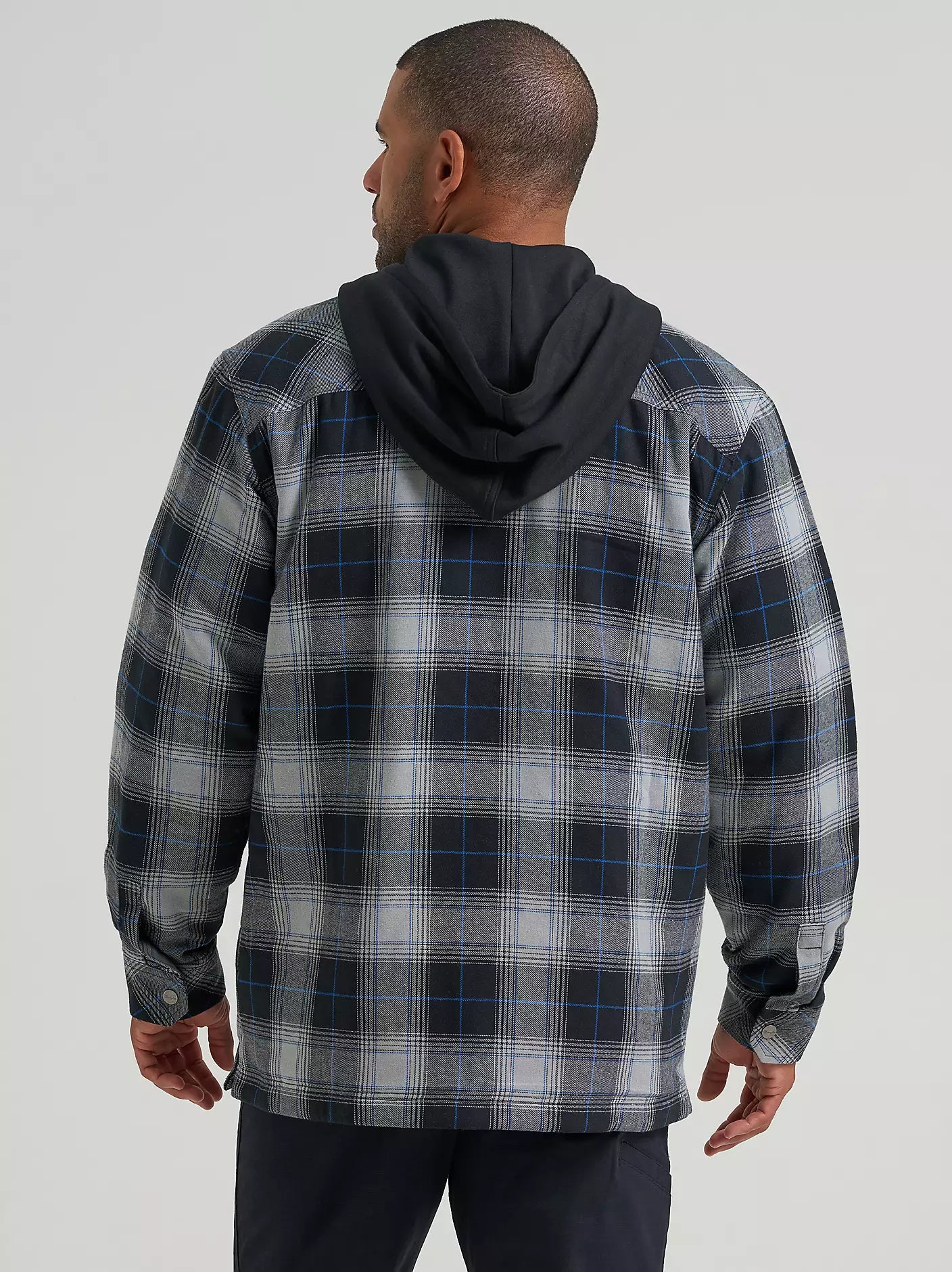 A man wearing a long-sleeve plaid hooded Wrangler Jacket in gray, black, and blue, paired with a black t-shirt and dark pants, against a solid light gray background.