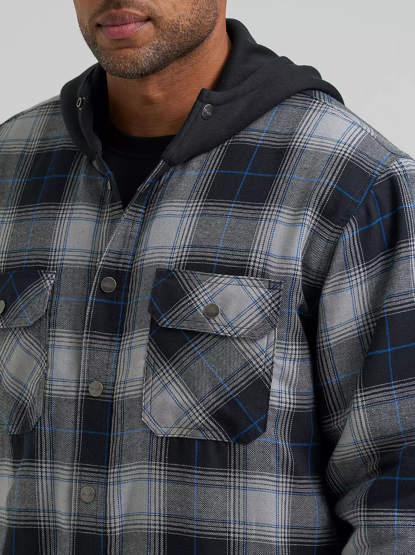 A close-up of a man wearing a gray and black plaid hooded Wrangler Jacker with blue line details, layered over a black hoodie and black t-shirt, against a solid gray background.