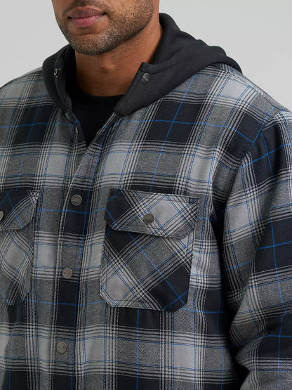 A close-up of a man wearing a gray and black plaid hooded Wrangler Jacker with blue line details, layered over a black hoodie and black t-shirt, against a solid gray background.