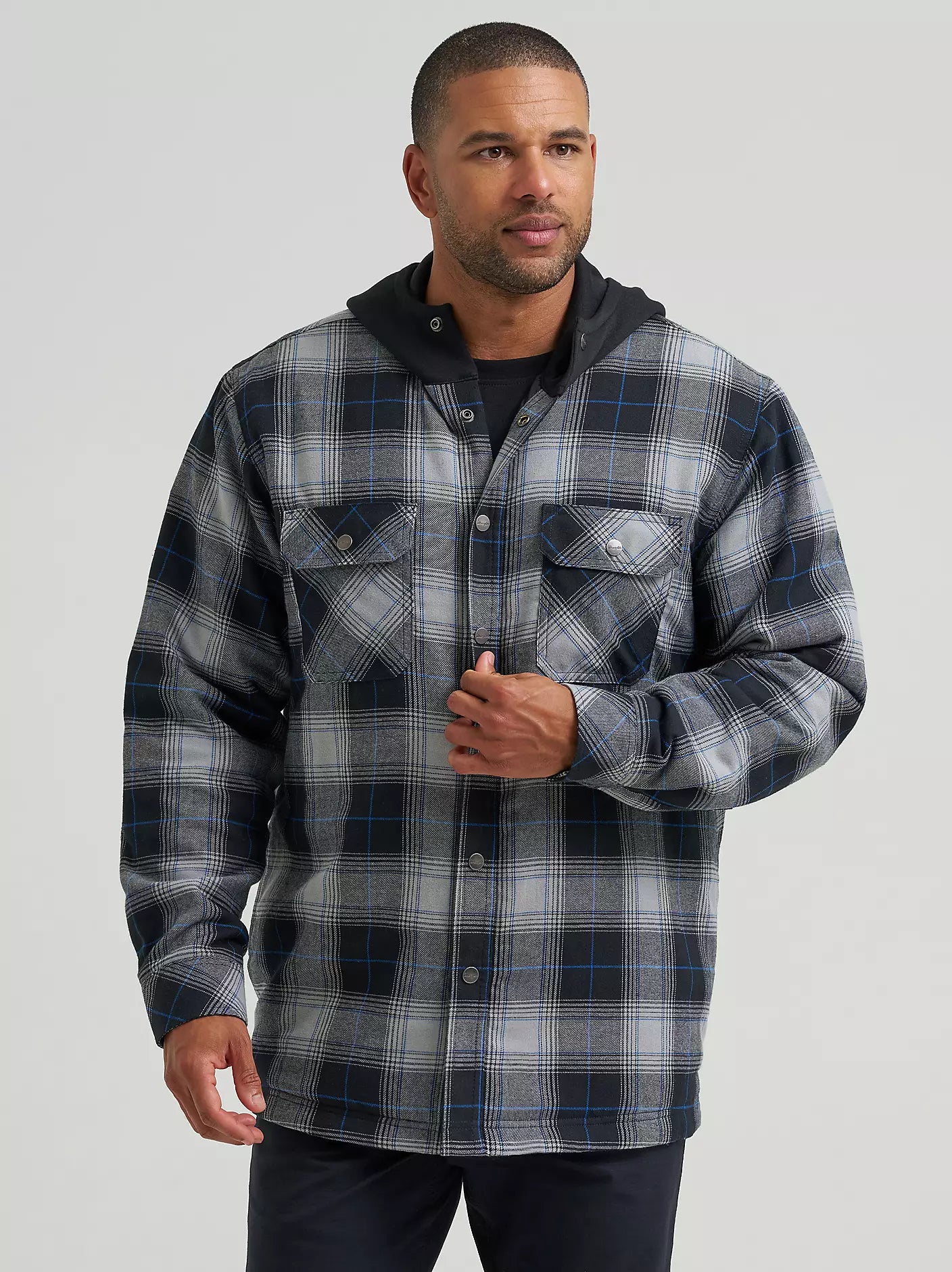 A man wearing a long-sleeve plaid hooded Wrangler Jacket in gray, black, and blue, paired with a black t-shirt and dark pants, against a solid light gray background.
