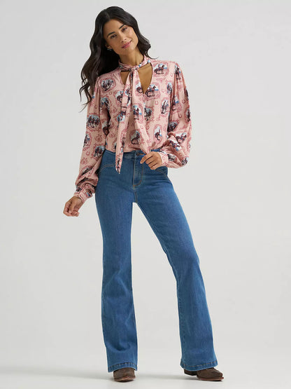 A woman modeling a pink long-sleeve blouse from Wrangler with a black and white graphic print, featuring a keyhole tie neckline, paired with blue flare jeans and brown shoes, against a light gray studio background.