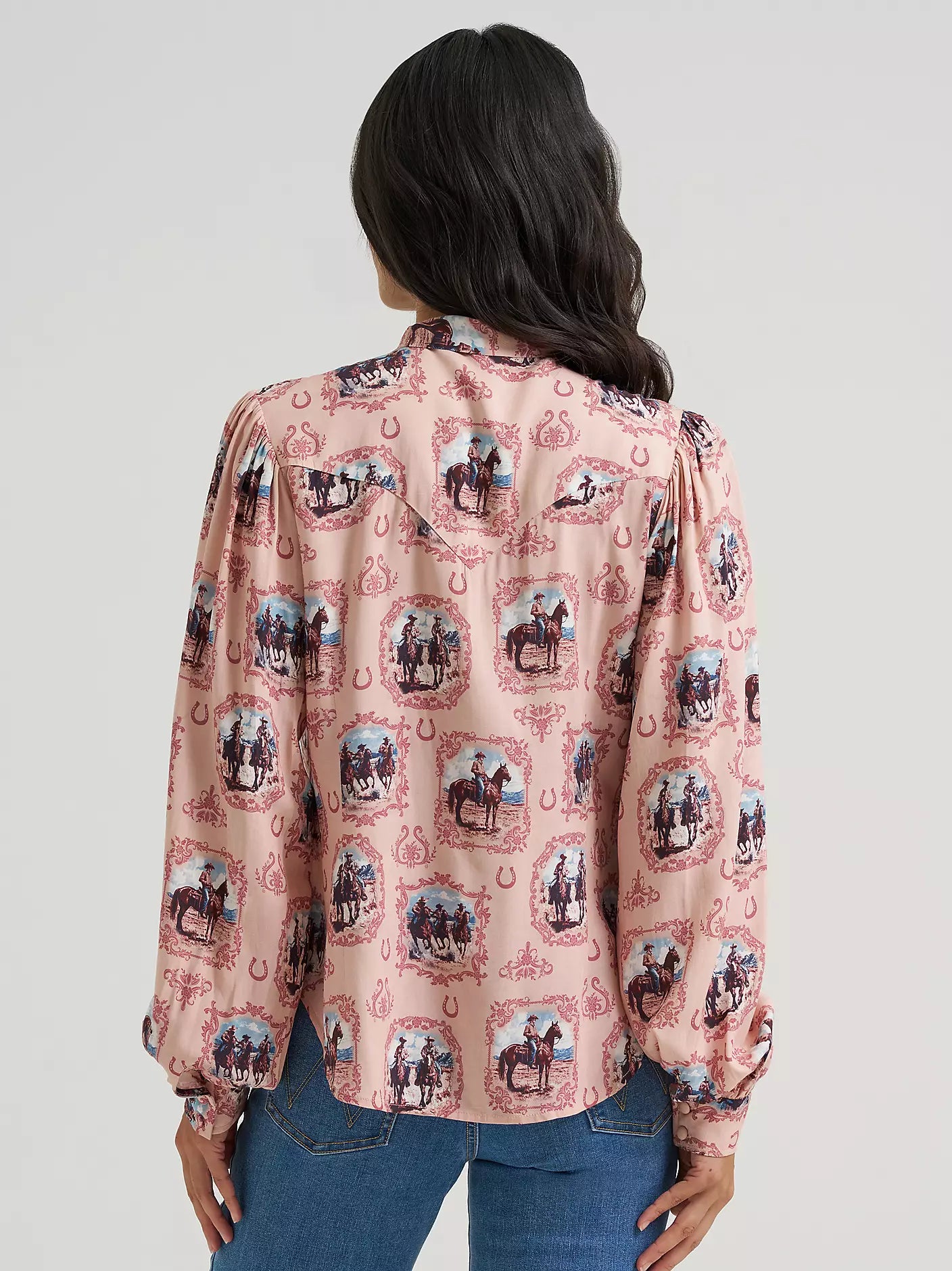 A woman viewed from the back wearing a pink long-sleeve blouse from Wrangler with an allover western-themed print of horseshoes and riders, paired with blue jeans against a light gray studio background.
