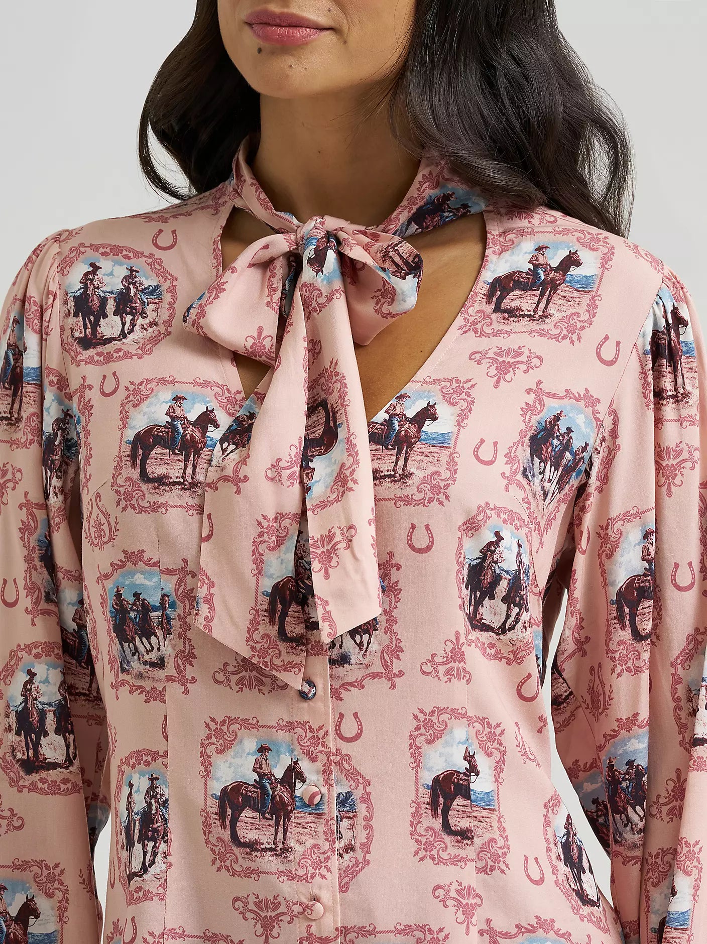 A close-up of a woman wearing a light pink long-sleeve blouse from Wrangler with a tie-neck detail and an allover western-themed print featuring horses, riders, horseshoes, and decorative frames.