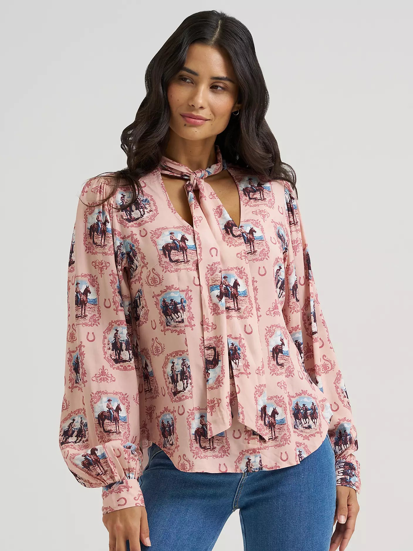 A woman modeling a pink long-sleeve blouse from Wrangler with a western-themed horse and rider print, featuring a tie-neck detail and front keyhole cutout, paired with blue jeans against a light gray studio background.