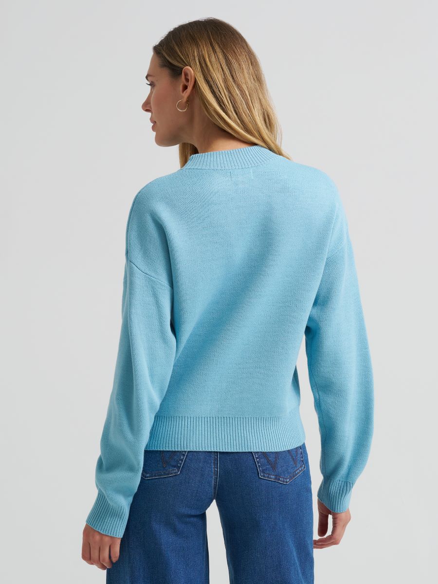 A woman shown from the back wearing a light blue Wrangler knit sweater and blue jeans against a plain light gray studio background.