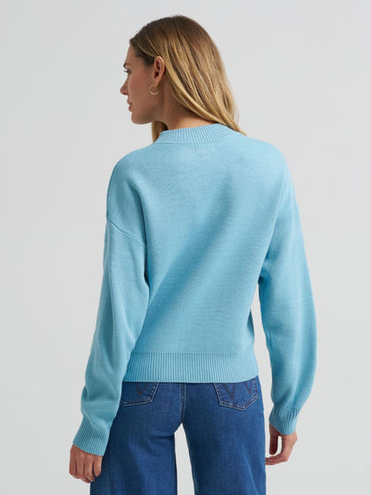 A woman shown from the back wearing a light blue Wrangler knit sweater and blue jeans against a plain light gray studio background.
