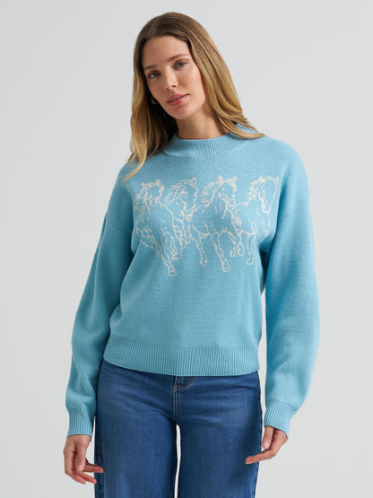 A woman wearing a light blue Wrangler crewneck sweater with a white embroidered horse pattern across the chest, paired with blue jeans, against a plain light gray studio background.