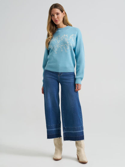 A woman modeling a light blue Wrangler crewneck sweater with a white embroidered mountain design, paired with wide-leg blue jeans and beige ankle boots, against a light gray studio background.