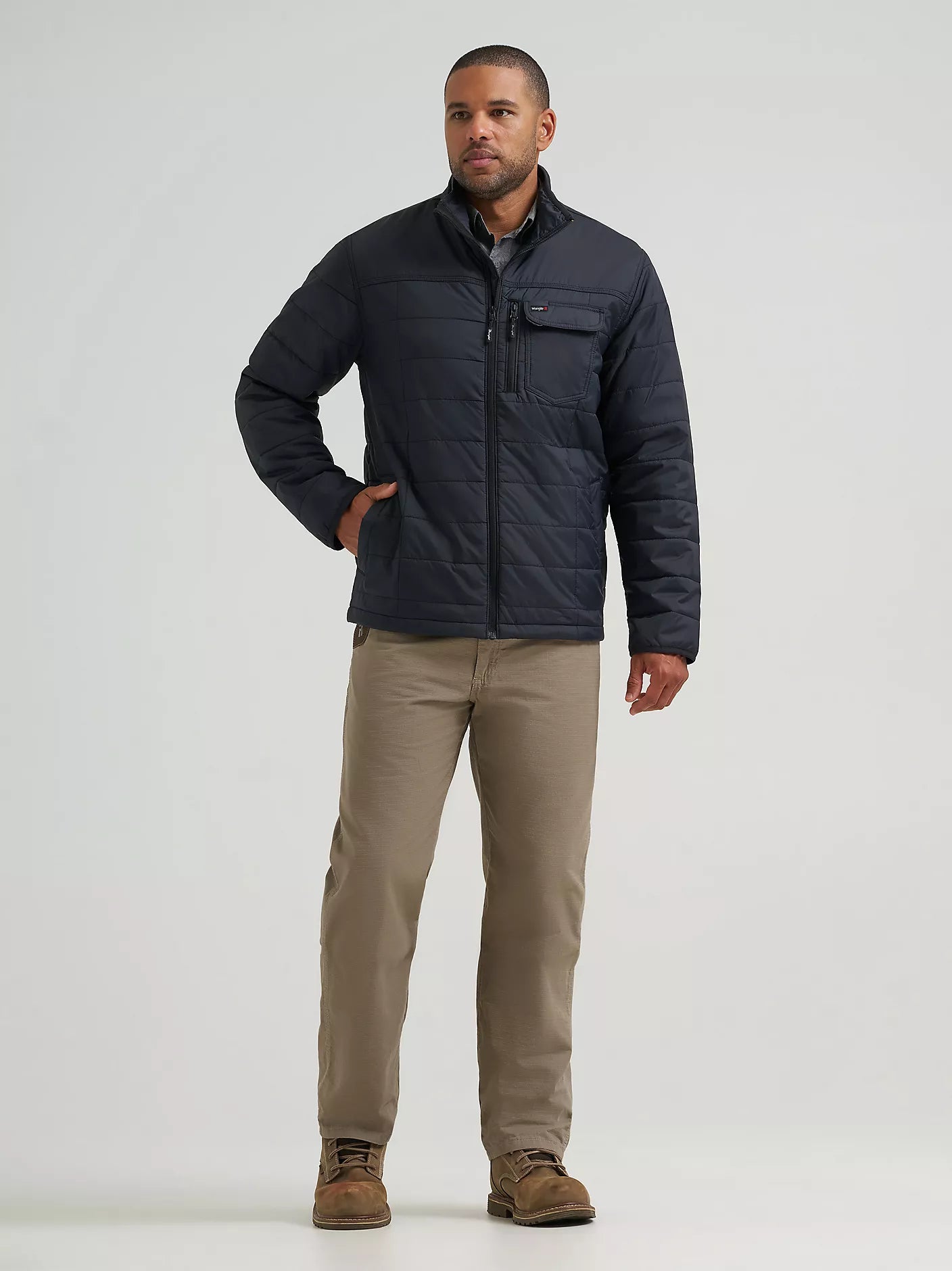 A man modeling a black Wrangler quilted jacket, paired with beige pants and brown shoes, standing against a light gray studio background.