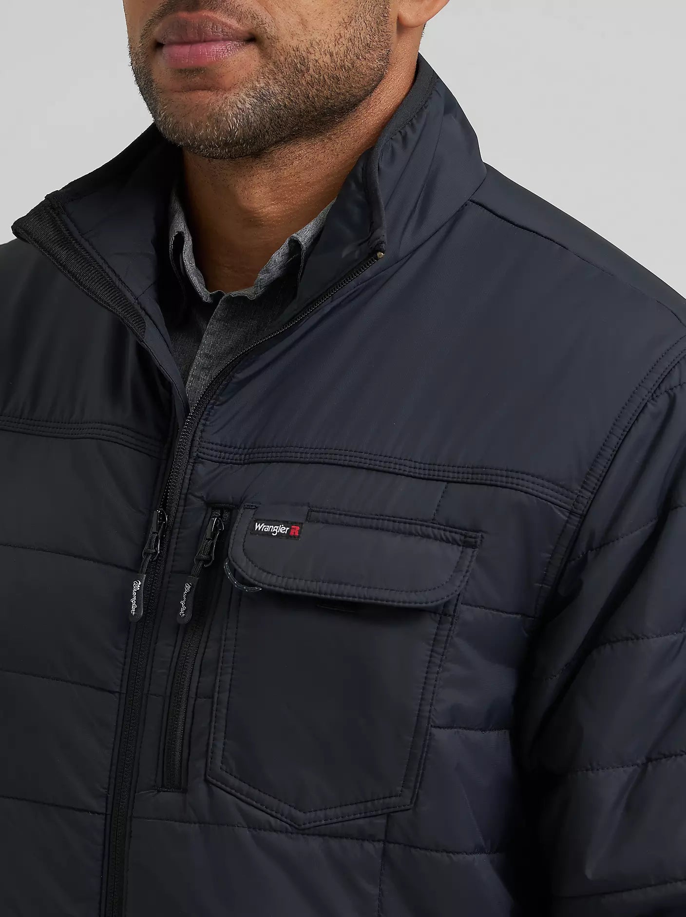 Close-up of a man wearing a black Wrangler quilted jacket with a zippered chest pocket and a small  logo, over a gray collared shirt.