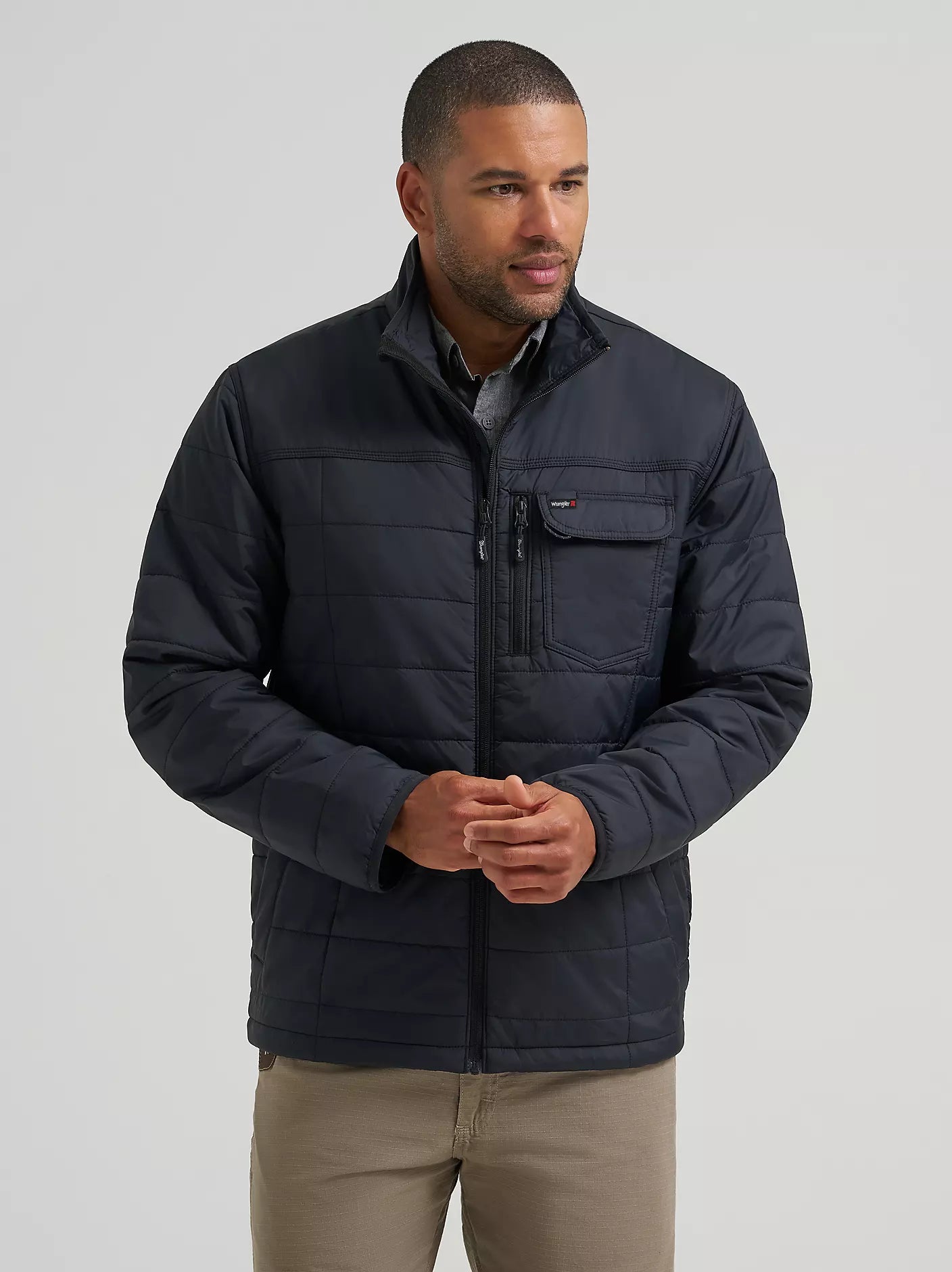 A man wearing a black Wrangler quilted jacket with a zippered chest pocket, standing against a light gray studio background.