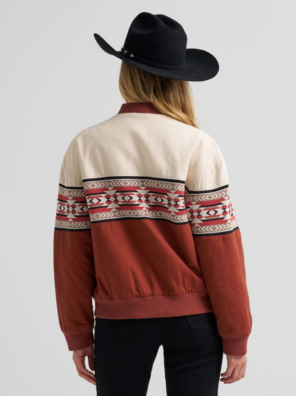 Woman wearing a orange and cream Wrangler bomber jacket, black jeans and a black cowboy hat on a grey background