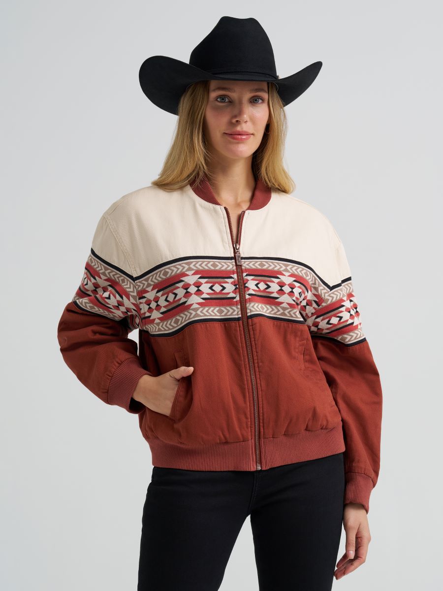 Woman wearing a orange and cream Wrangler bomber jacket, black jeans and a black cowboy hat on a grey background