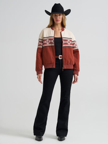 Woman wearing a orange and cream Wrangler bomber jacket, black jeans, boots and a black cowboy hat on a grey background