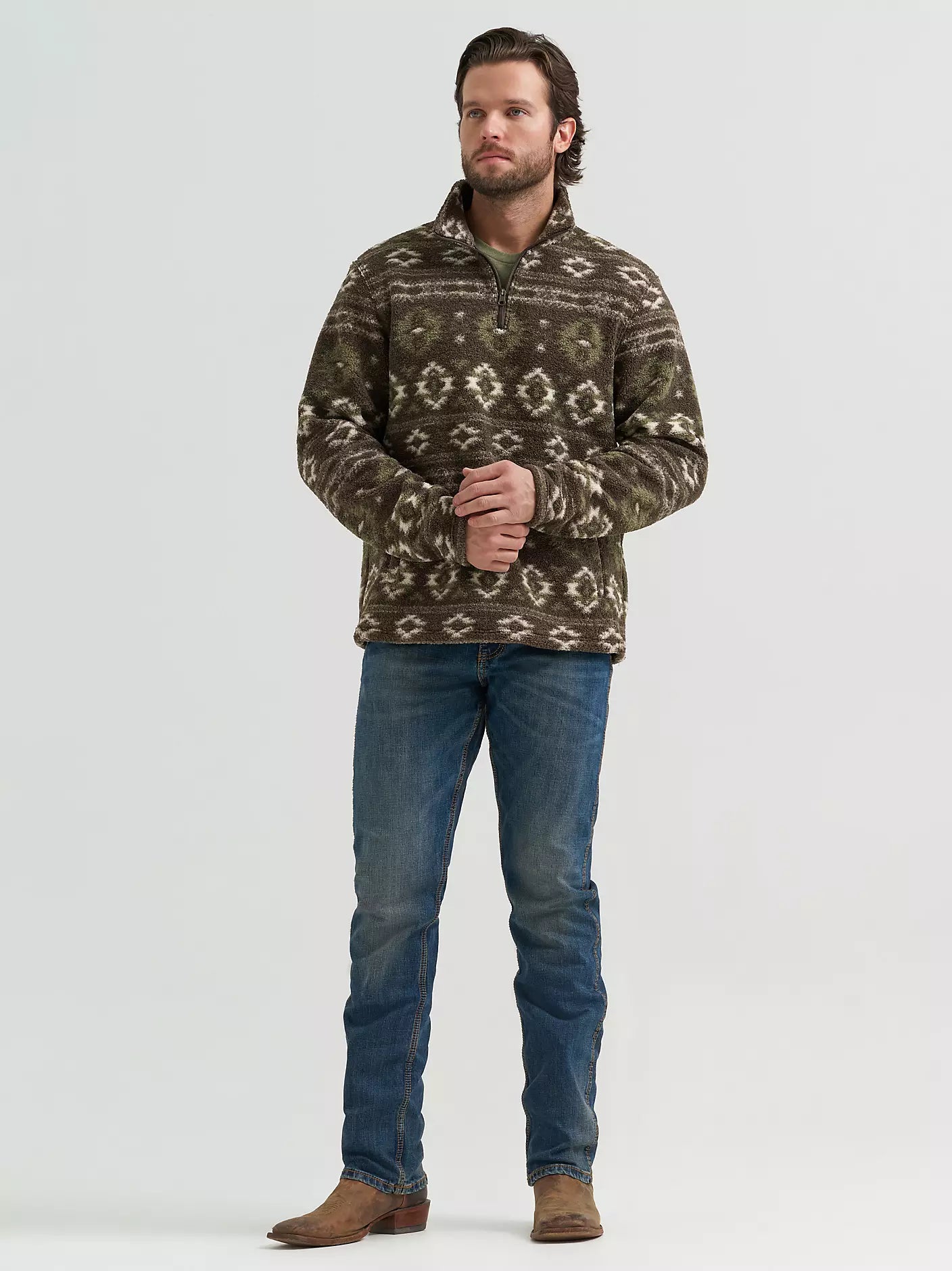 A man modeling a quarter-zip fleece pullover sweater from Wrangler with a brown, beige, and green geometric pattern, paired with blue jeans and brown boots, against a light gray studio background.