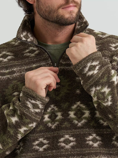 A close-up of a man wearing a brown and olive green patterned fleece quarter-zip pullover, from Wrangler, pulling the zipper with both hands. The background is a clean, neutral gray studio backdrop.