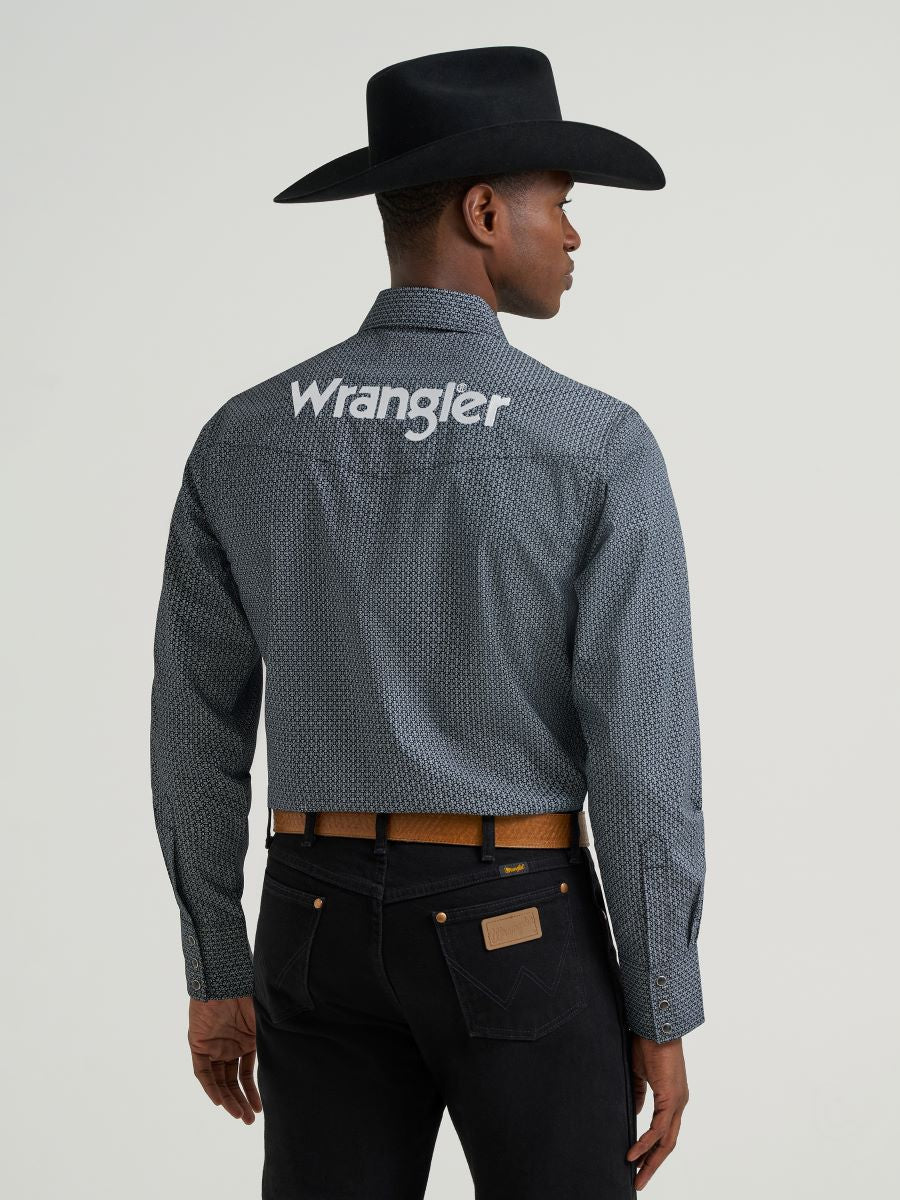 Man wearing a grey printed Wrangler shirt, black jeans and belt and a black cowboy hat on a grey background
