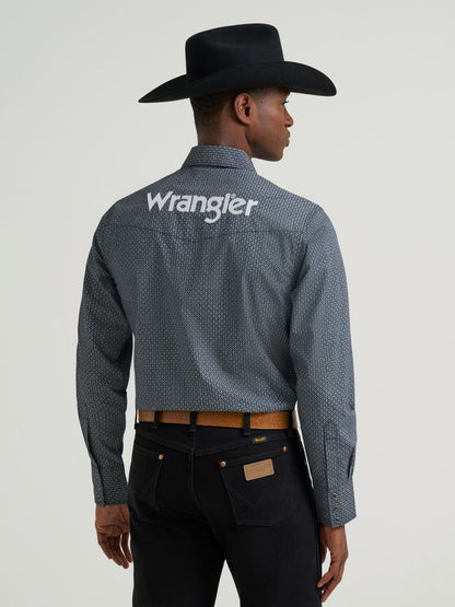 Man wearing a grey printed Wrangler shirt, black jeans and belt and a black cowboy hat on a grey background