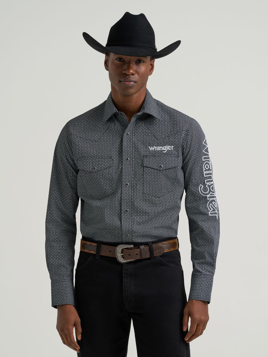 Man wearing a grey printed Wrangler shirt, black jeans and belt and a black cowboy hat on a grey background