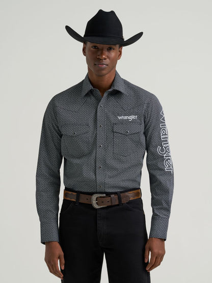 Man wearing a grey printed Wrangler shirt, black jeans and belt and a black cowboy hat on a grey background