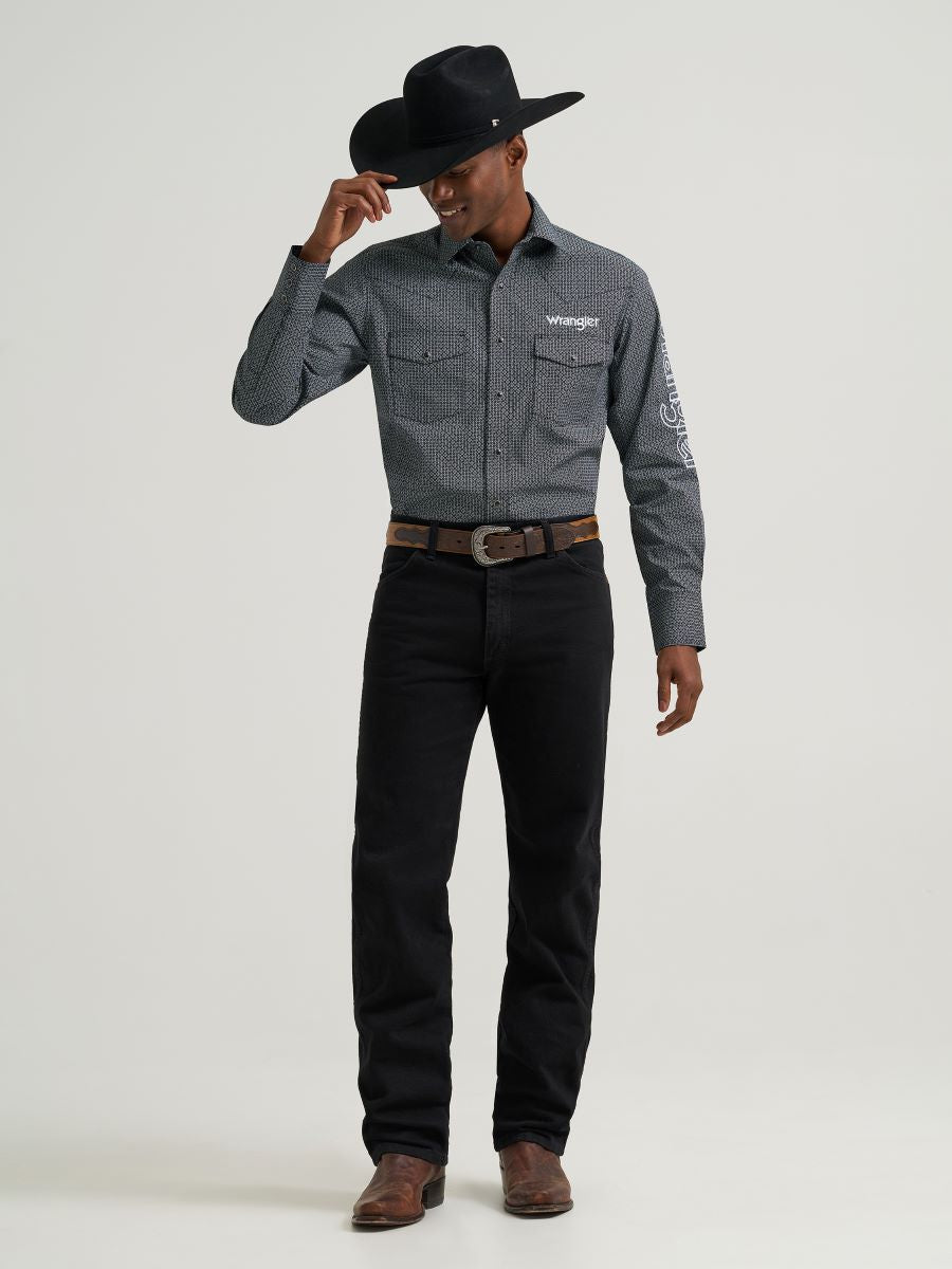 Man wearing a grey printed Wrangler shirt, black jeans and belt, boots and a black cowboy hat on a grey background