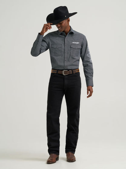 Man wearing a grey printed Wrangler shirt, black jeans and belt, boots and a black cowboy hat on a grey background