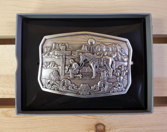 A Praying Desert Cowboy Belt Buckle by Nocona, displayed in its open box, showing a silver belt buckle with an engraved scene of a cowboy and a horse in a desert landscape.