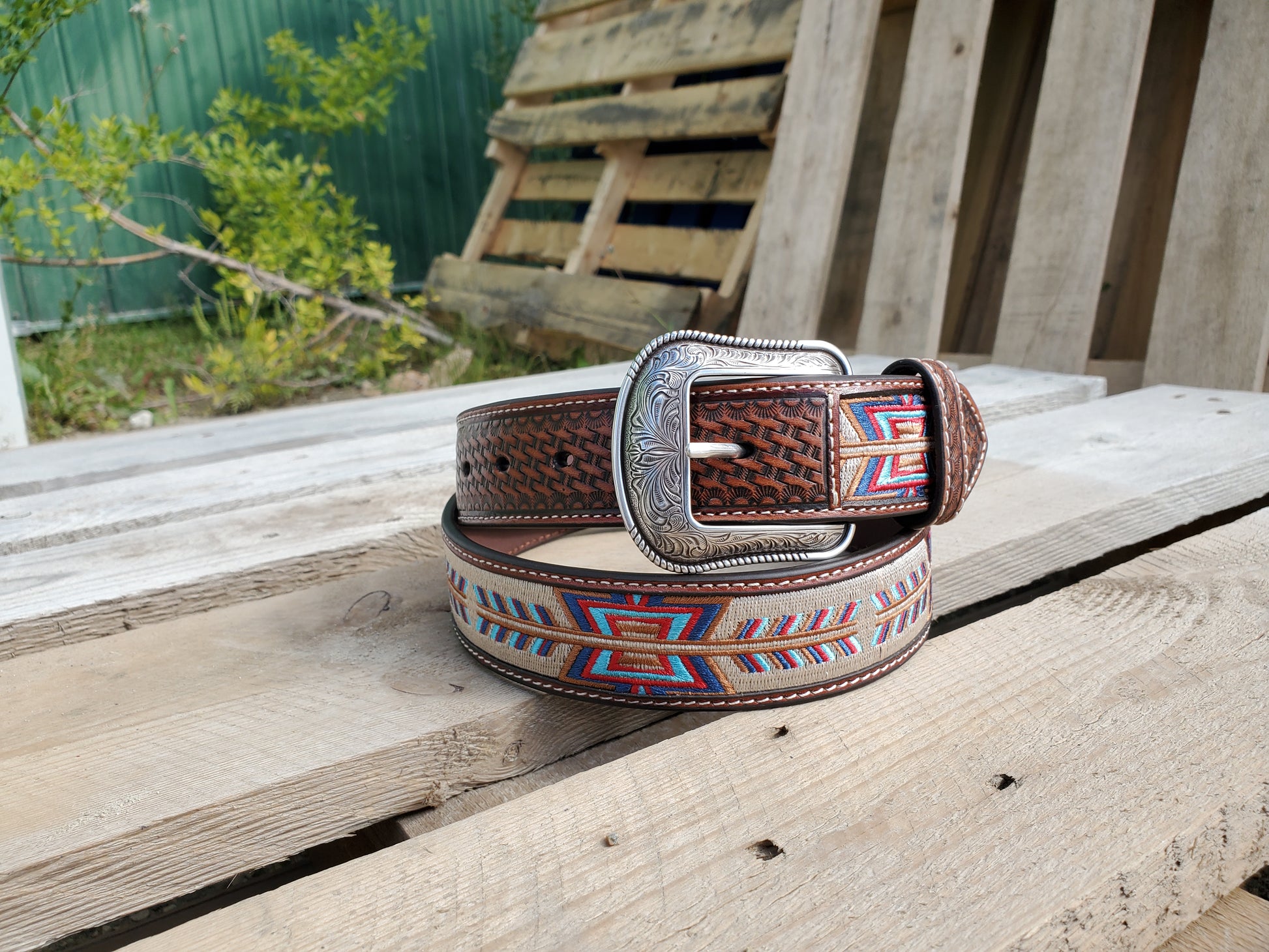 A brown leather belt with 'Fletching' stitch embroidery and basket weave detail, displayed on a wooden surface with a belt buckle