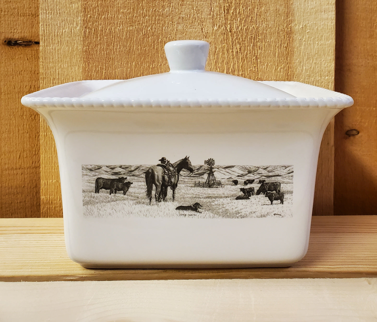 A white ceramic butter dish with a lid, featuring a black and white illustration of a man and his horse looking at cows in a pasture