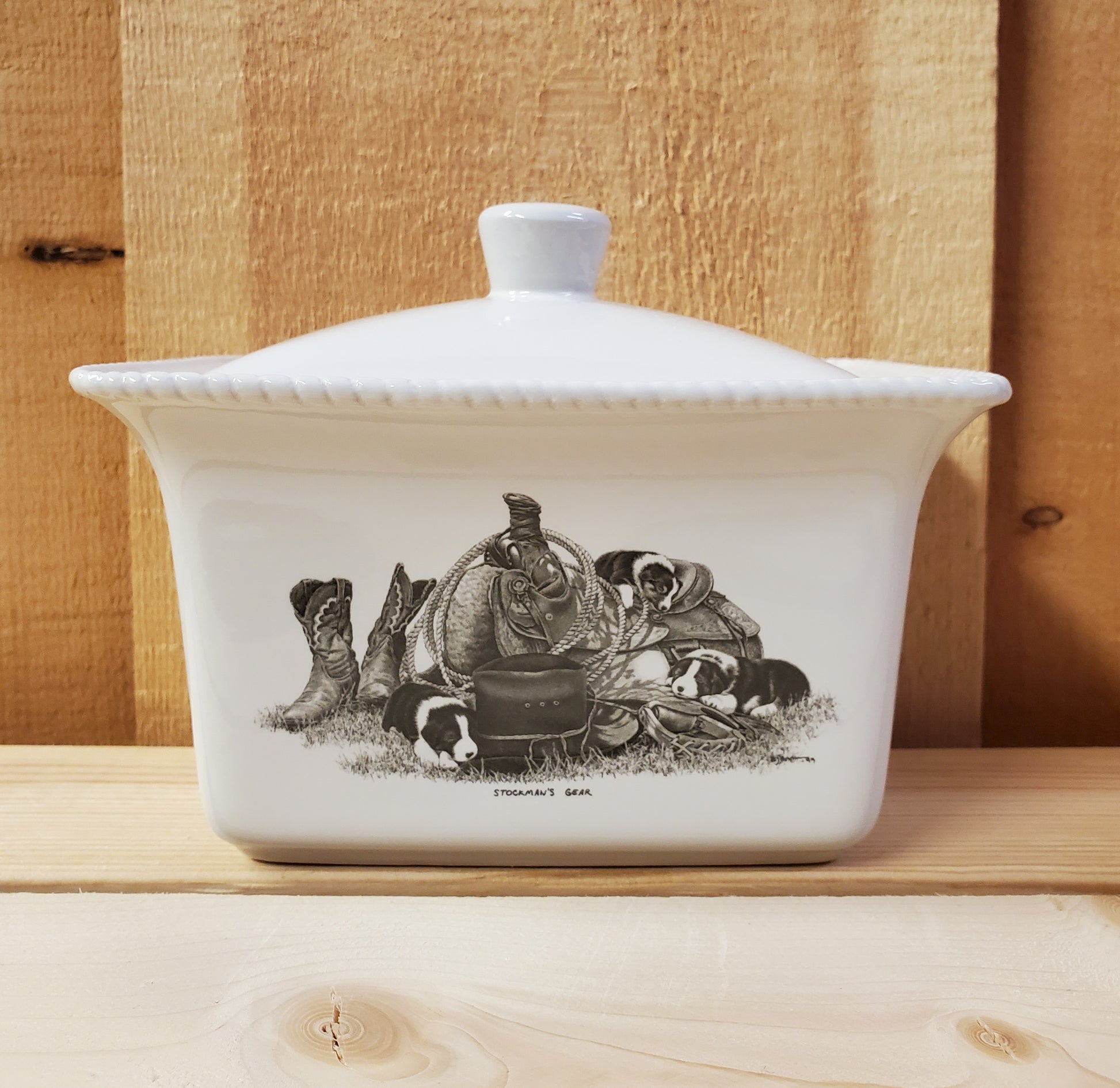 A white ceramic butter dish with a lid, featuring a black and white illustration of puppies on a western saddle
