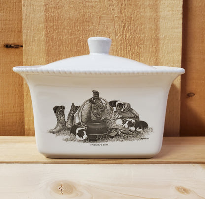 A white ceramic butter dish with a lid, featuring a black and white illustration of puppies on a western saddle