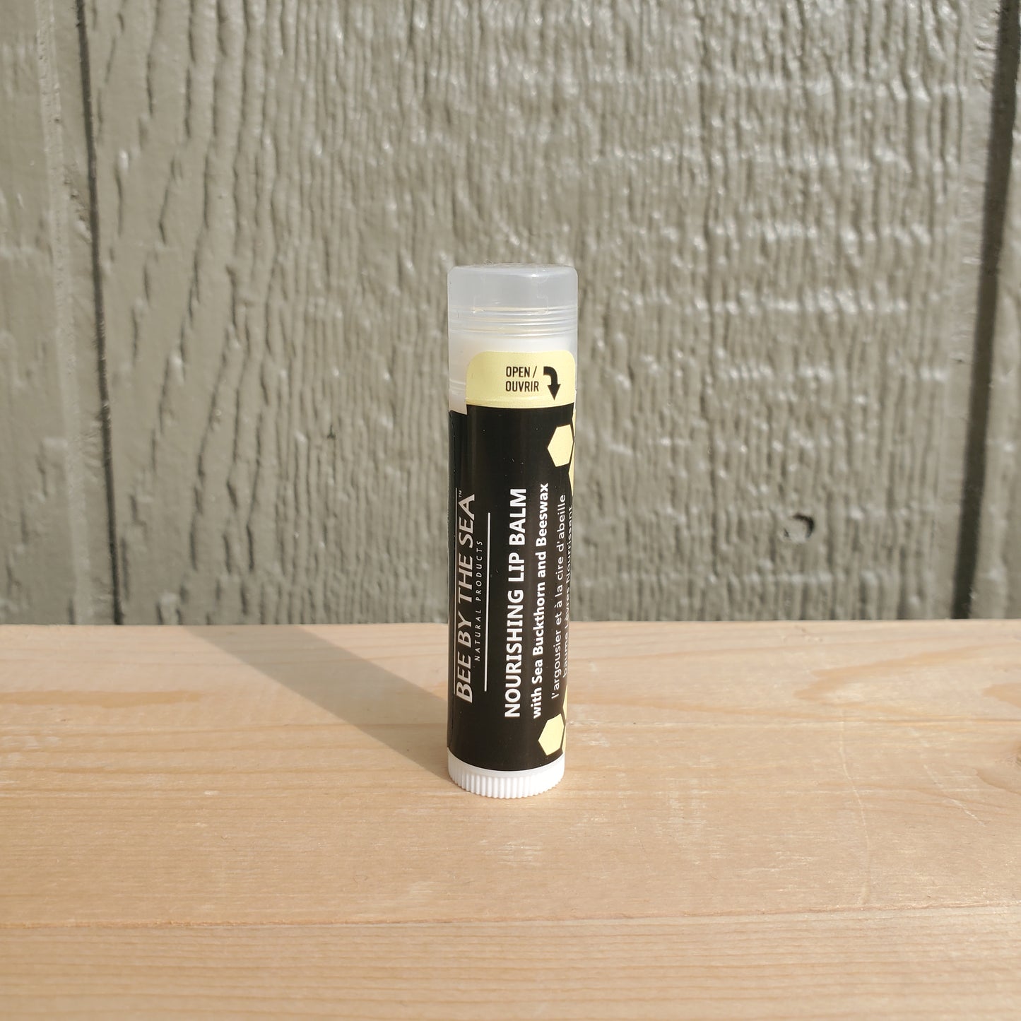A tube of Bee By The Sea Nourishing Lip Balm placed upright on a wooden surface with a plain background.