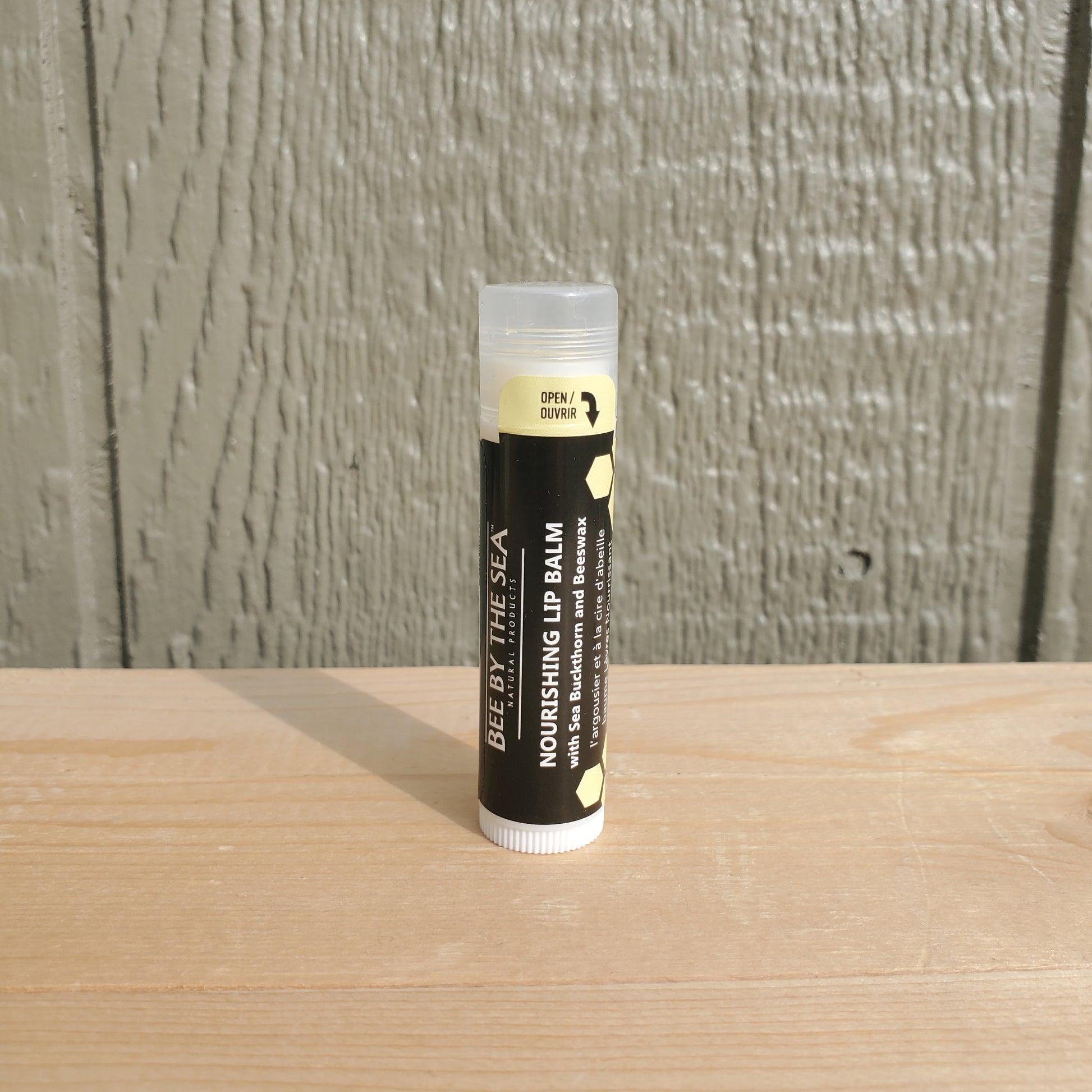 A tube of Bee By The Sea Nourishing Lip Balm placed upright on a wooden surface with a plain background.