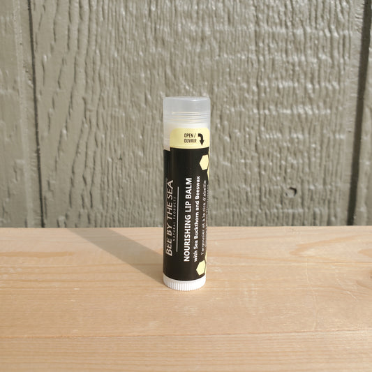 A tube of Bee By The Sea Nourishing Lip Balm placed upright on a wooden surface with a plain background.
