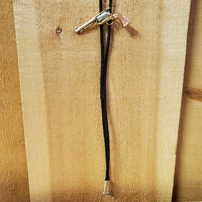 A tri-tone revolver bolo tie with a silver body and a brown barrel, attached to a black rope and a wooden surface.