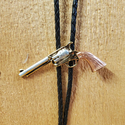 A tri-tone revolver bolo tie with a silver body and a brown barrel, attached to a black rope and a wooden surface.